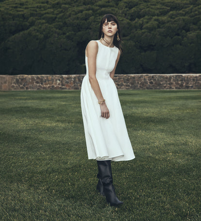 Woman in a white dress and black boots standing on grass with a blurred background.