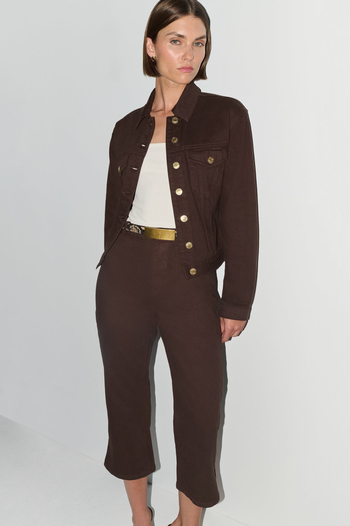 A model wearing a dark brown cropped jacket and matching cropped trousers with gold-tone buttons and a gold belt, styled over a white top against a light gray studio background.