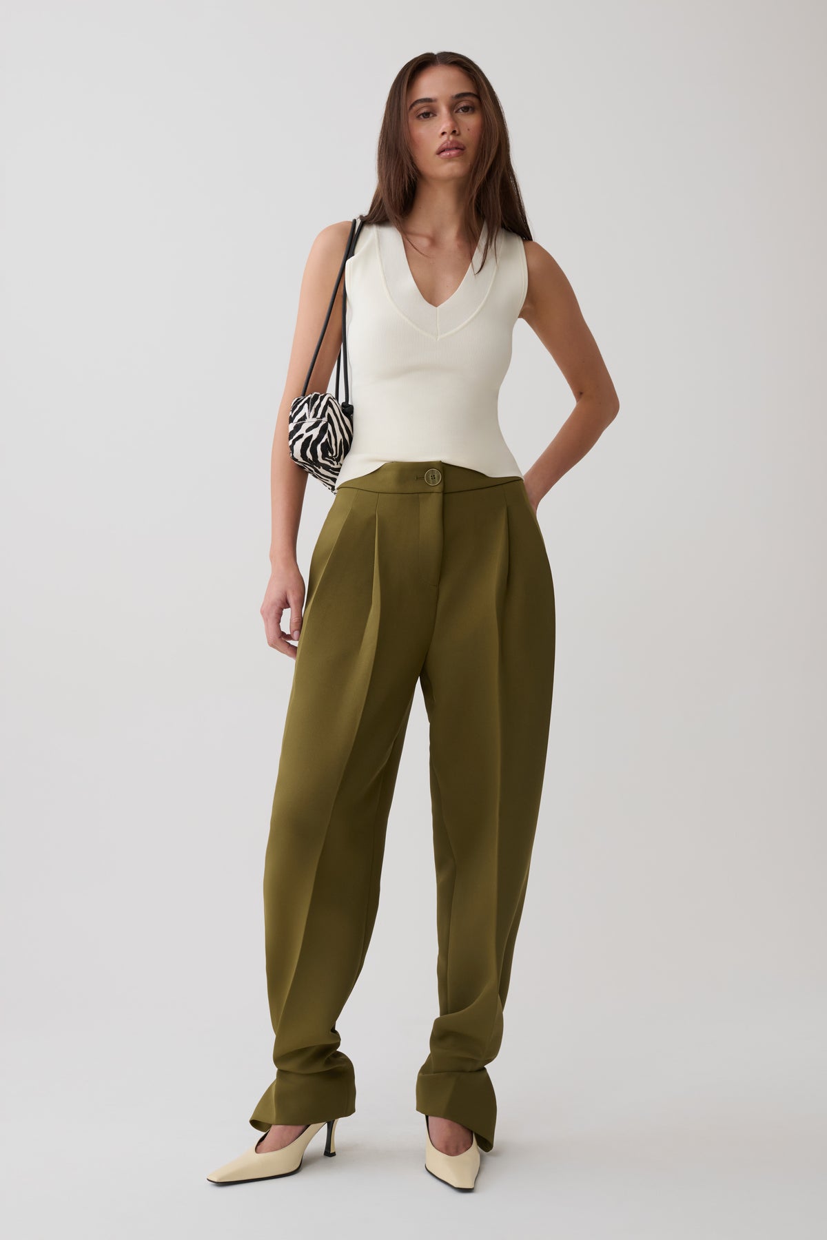 A model stands against a light gray studio background wearing olive green wide-leg trousers, a white sleeveless top, and cream pointed-toe heels, while carrying a black-and-white patterned shoulder bag.