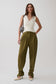 A model stands against a light gray studio background wearing olive green wide-leg trousers, a white sleeveless top, and cream pointed-toe heels, while carrying a black-and-white patterned shoulder bag.