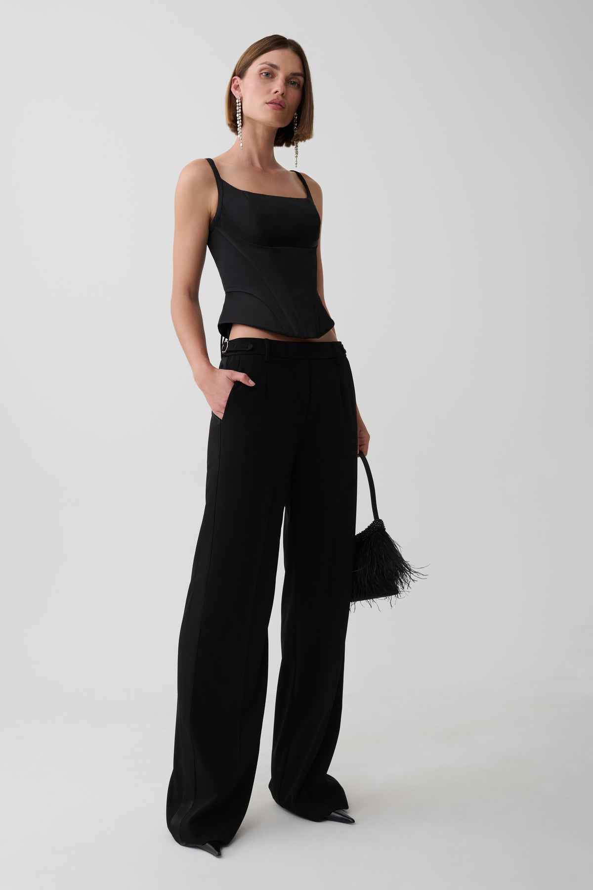 A model stands against a light gray studio background wearing a black sleeveless top, black wide-leg trousers, and holding a small black feather-trim handbag.