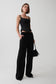 A model stands against a light gray studio background wearing a black sleeveless top, black wide-leg trousers, and holding a small black feather-trim handbag.