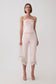 A model stands against a light gray studio background wearing a pale pink strapless top with ruffled detailing and matching cropped pants with floral lace trim at the hems. She is holding a small cream handbag and wearing clear high-heel sandals.
