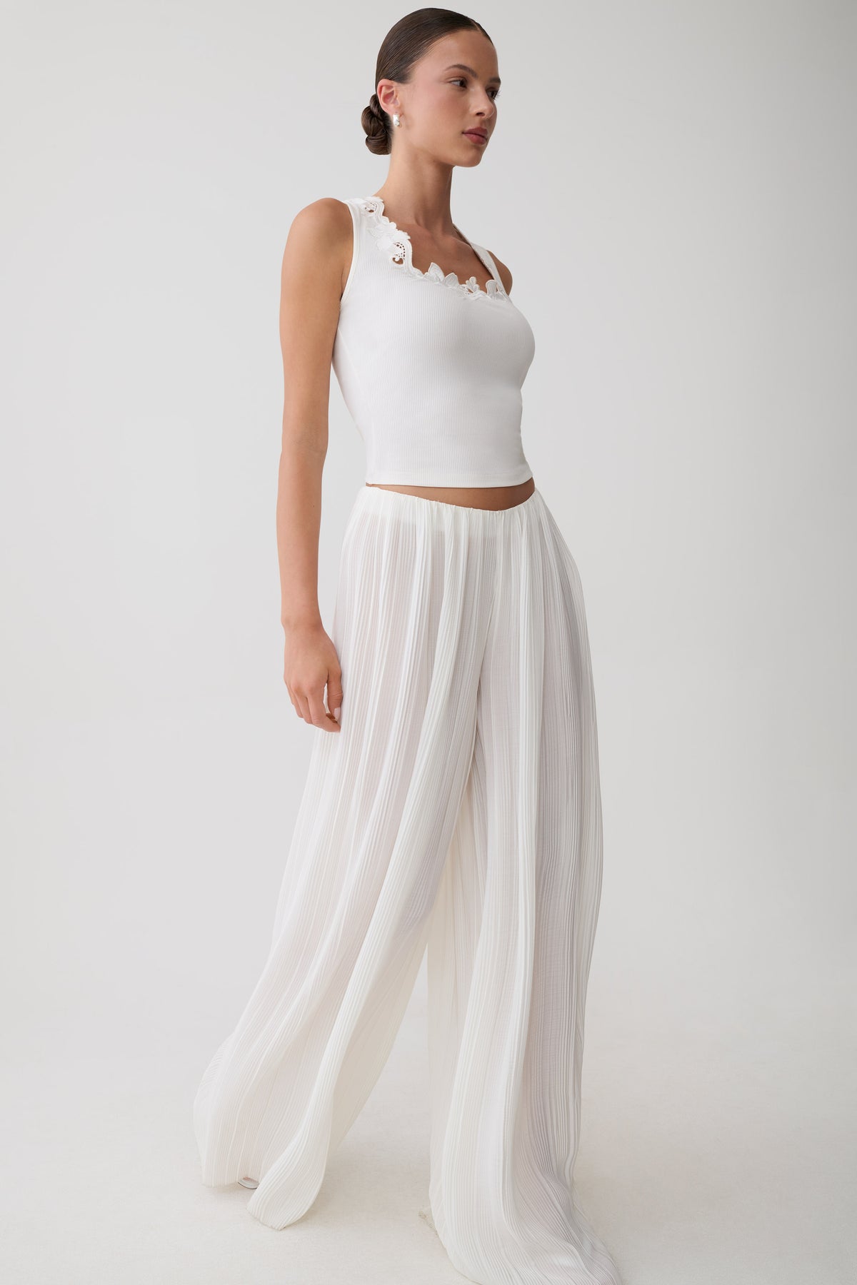 A woman modeling a white sleeveless crop top with scalloped trim and matching white wide-leg pleated pants against a plain light gray studio background.