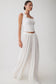A woman modeling a white sleeveless crop top with scalloped trim and matching white wide-leg pleated pants against a plain light gray studio background.