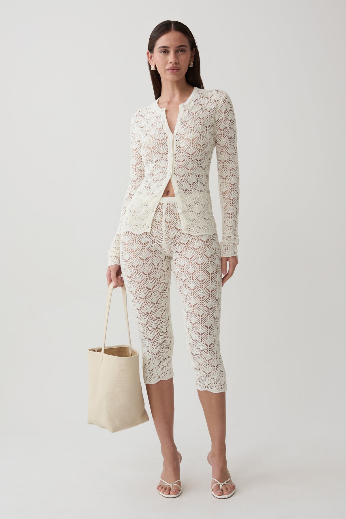 A model stands against a light gray studio background wearing a matching cream lace three-piece set consisting of a long-sleeve button-front cardigan, cropped capri pants, and a tie belt. She is holding a cream canvas tote bag and wearing strappy high-heeled sandals.