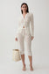 A model stands against a light gray studio background wearing a matching cream lace three-piece set consisting of a long-sleeve button-front cardigan, cropped capri pants, and a tie belt. She is holding a cream canvas tote bag and wearing strappy high-heeled sandals.