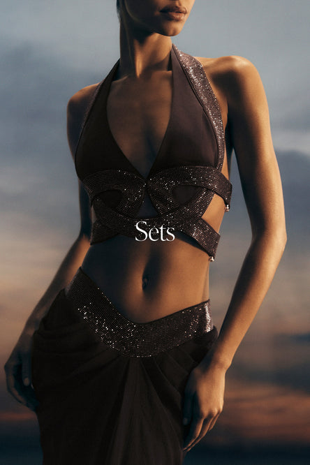 Model wearing a dark, embellished outfit with 'Sets' branding against a blurred natural background.