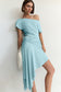 A woman models an off-the-shoulder light blue dress with a draped, gathered skirt and asymmetrical hem, holding a white feathered clutch against a plain off-white wall.