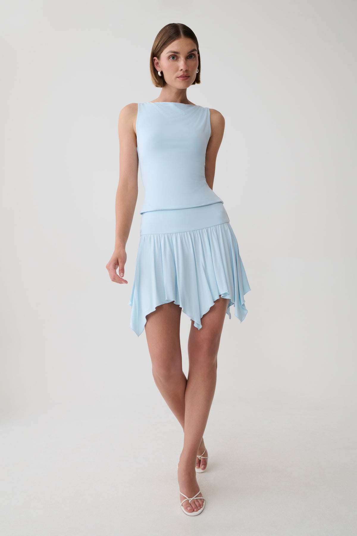 A woman modeling a light blue sleeveless top with a fitted bodice and a pleated ruffled mini skirt, shown against a plain light gray studio background.