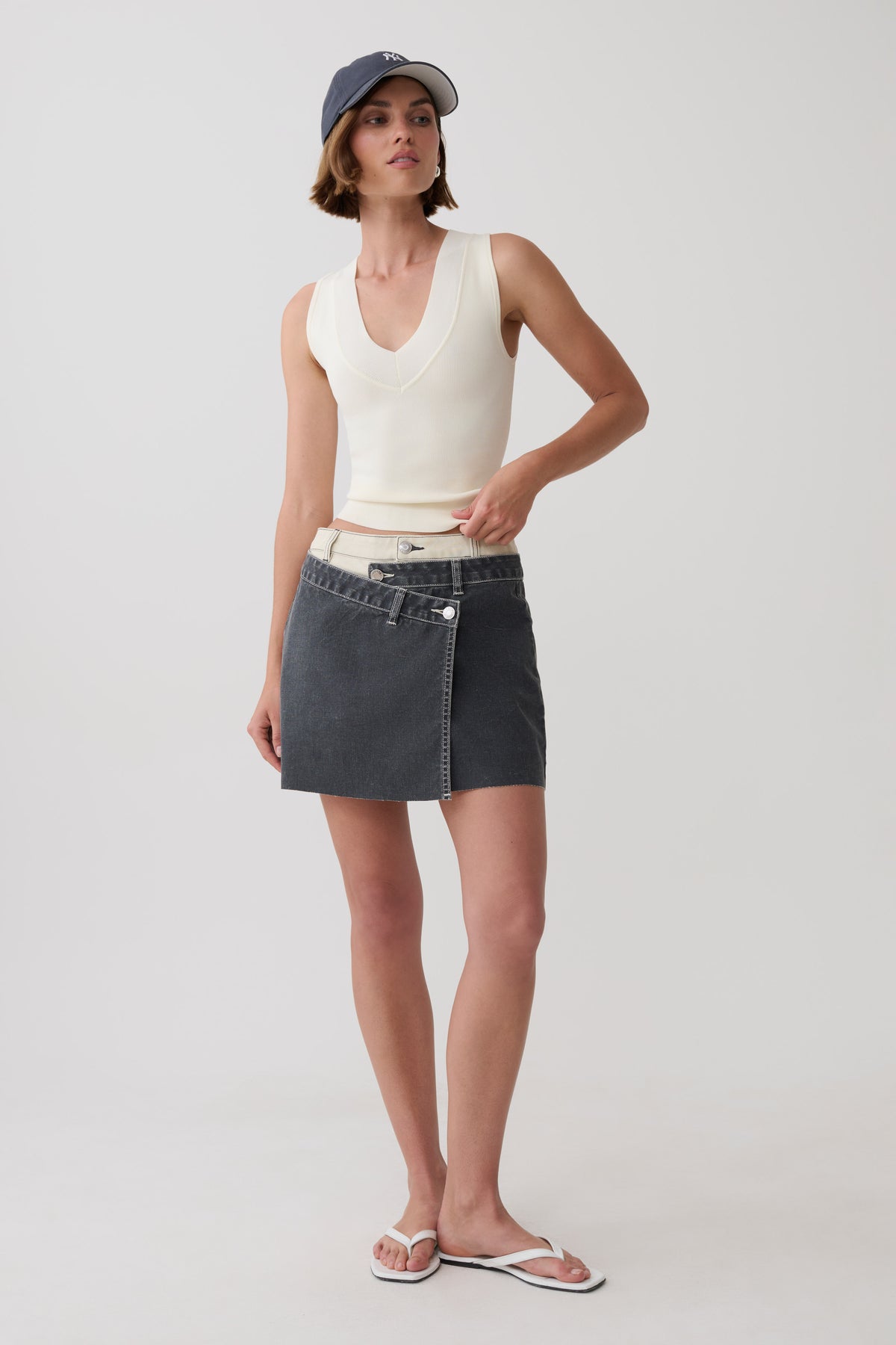 A model stands against a light gray studio background wearing a cream ribbed sleeveless crop top, a dark gray wrap-style denim mini skirt with a white waistband, a navy baseball cap, and white flip-flop sandals.