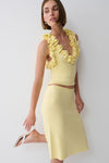 A woman models a pale yellow two-piece knit set consisting of a ruffled halter crop top and a matching midi skirt, paired with white heeled sandals against a plain light gray background.