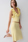 A woman models a pale yellow two-piece knit set consisting of a ruffled halter crop top and a matching midi skirt, paired with white heeled sandals against a plain light gray background.