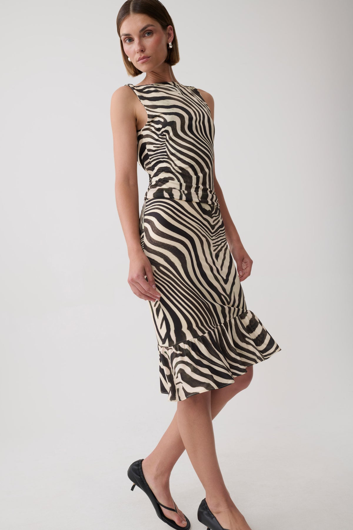 A woman modeling a sleeveless halter-neck midi dress with a bold black and beige zebra-like wavy stripe pattern, paired with black heels, against a plain light gray studio background.