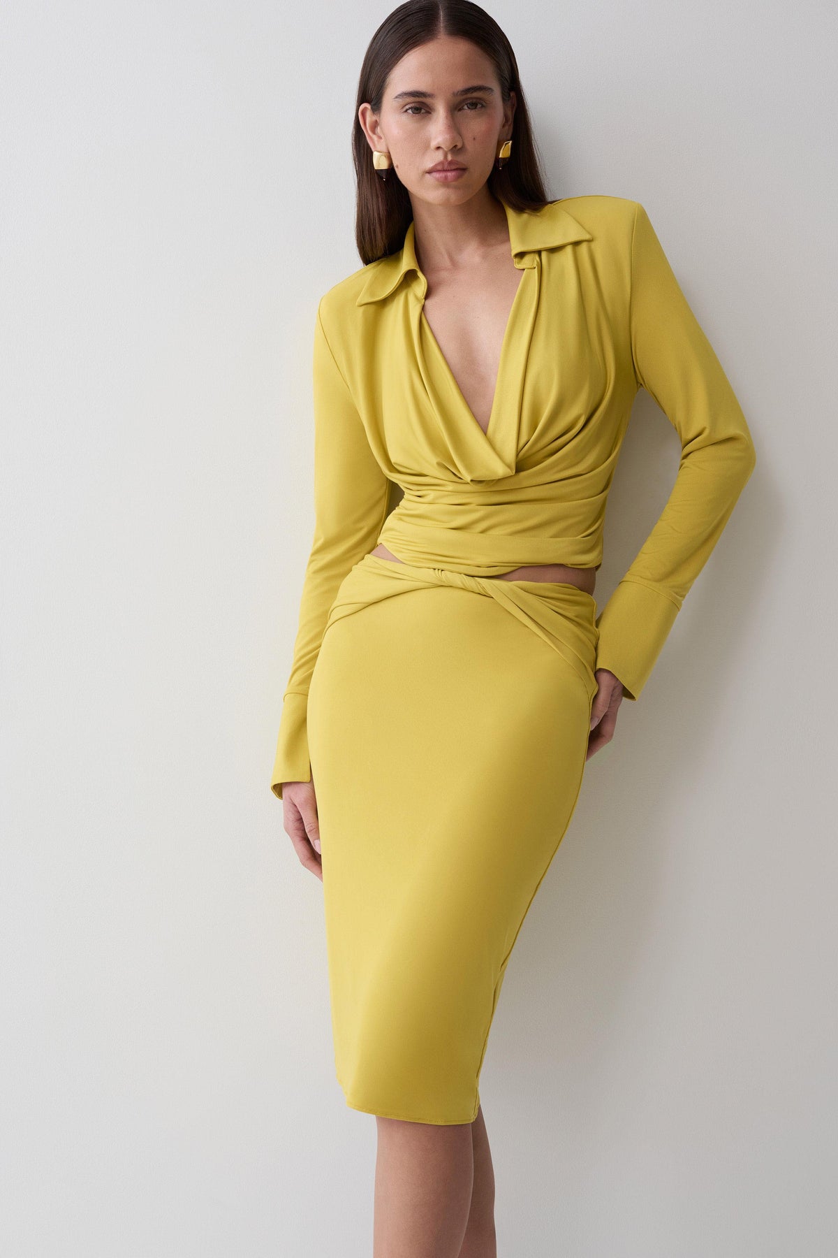 A woman modeling a bright yellow two-piece outfit consisting of a long-sleeve collared top with a twisted wrap detail at the waist and a matching fitted midi skirt, posed against a plain light gray wall.