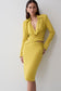 A woman modeling a bright yellow two-piece outfit consisting of a long-sleeve collared top with a twisted wrap detail at the waist and a matching fitted midi skirt, posed against a plain light gray wall.