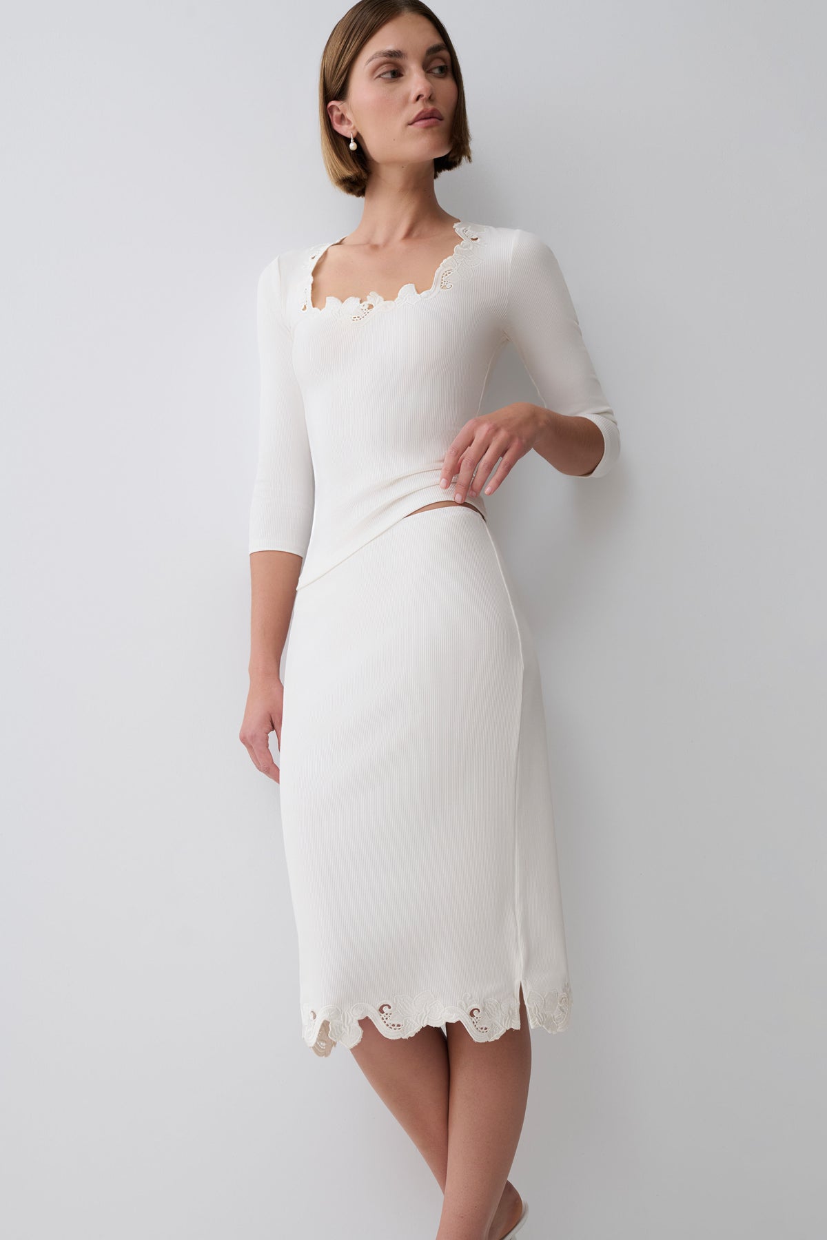 A woman modeling a white three-quarter sleeve top with scalloped eyelet trim at the neckline and hem paired with a matching midi skirt set against a plain light gray studio background.