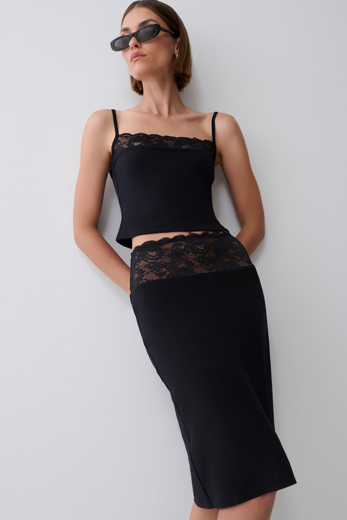 A woman modeling a black two-piece set consisting of a spaghetti-strap crop top with lace trim and a matching high-waisted pencil skirt with lace detailing, styled with dark sunglasses against a plain light gray wall.