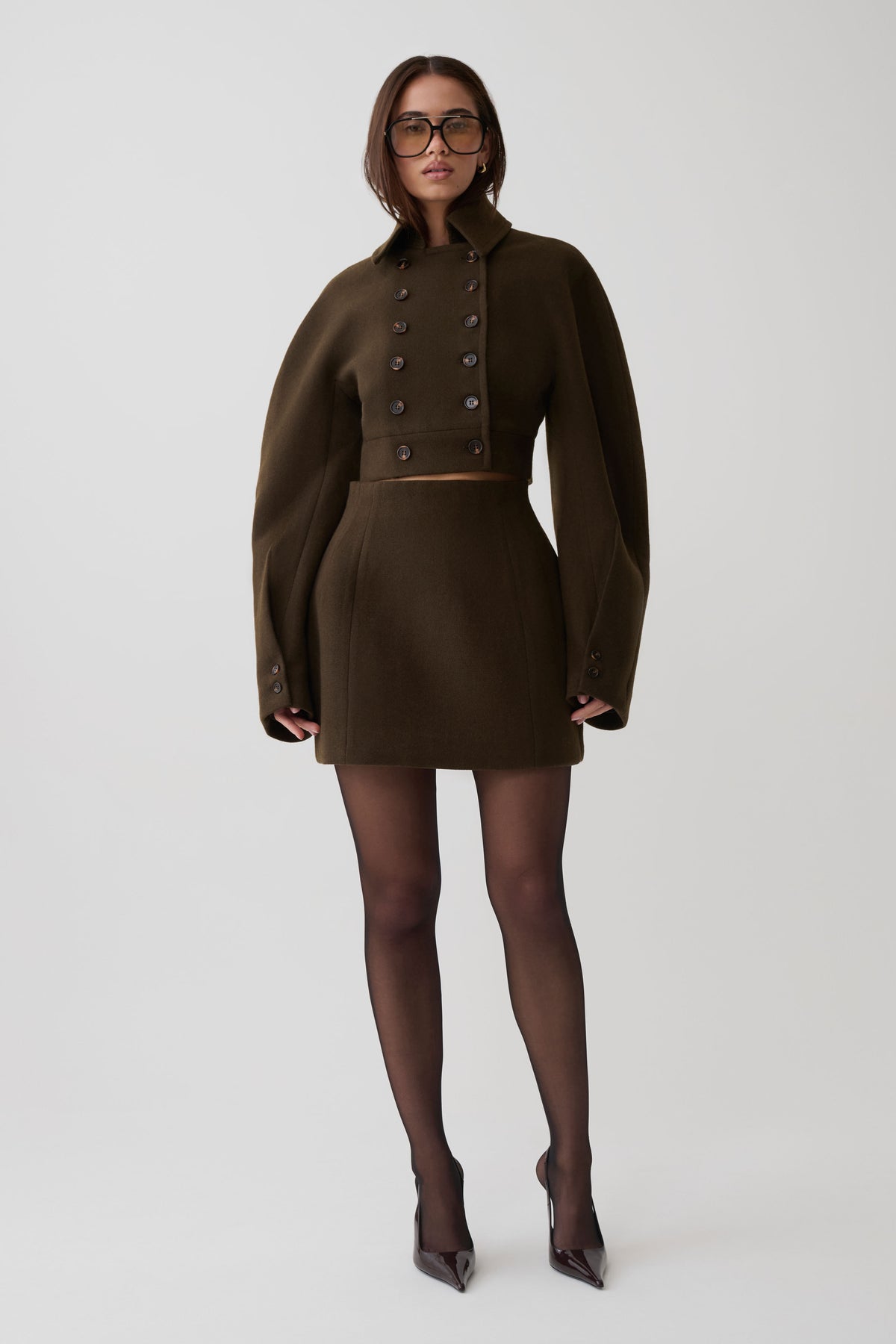 A model wearing a dark brown double-breasted mini coat with oversized sleeves, paired with sheer black tights and dark pointed-toe heels, standing against a light gray studio background.