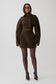 A model wearing a dark brown double-breasted mini coat with oversized sleeves, paired with sheer black tights and dark pointed-toe heels, standing against a light gray studio background.