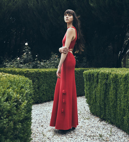 Woman in a red maxi dress standing in a garden with manicured hedges.