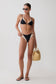 A model wearing a black and white triangle bikini, black sunglasses, and black flip-flops, holding a woven straw bag against a plain light gray studio background.