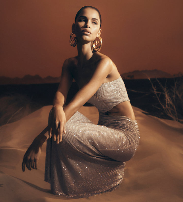 Woman in a sparkling dress sitting on sand with a warm-toned background.