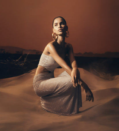 Woman in a sparkling dress sitting on sand dunes with a warm-toned sky.