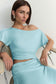 A woman wearing a light blue off-the-shoulder two-piece set with a cropped top and ruched skirt, seated on a chair against a plain light background.