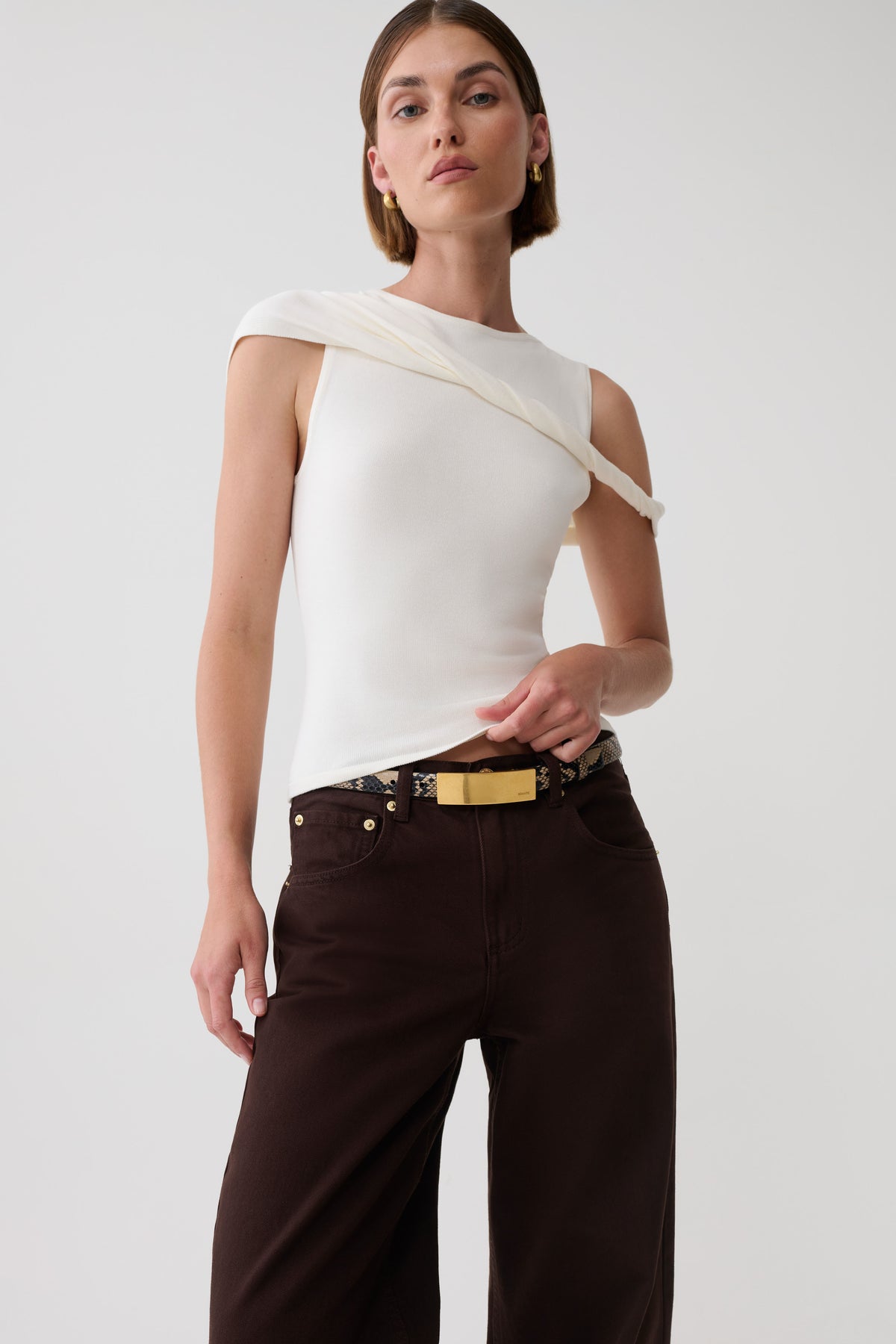 A model wearing a white asymmetrical draped top and dark brown wide-leg trousers with a gold belt, photographed against a light gray studio background.