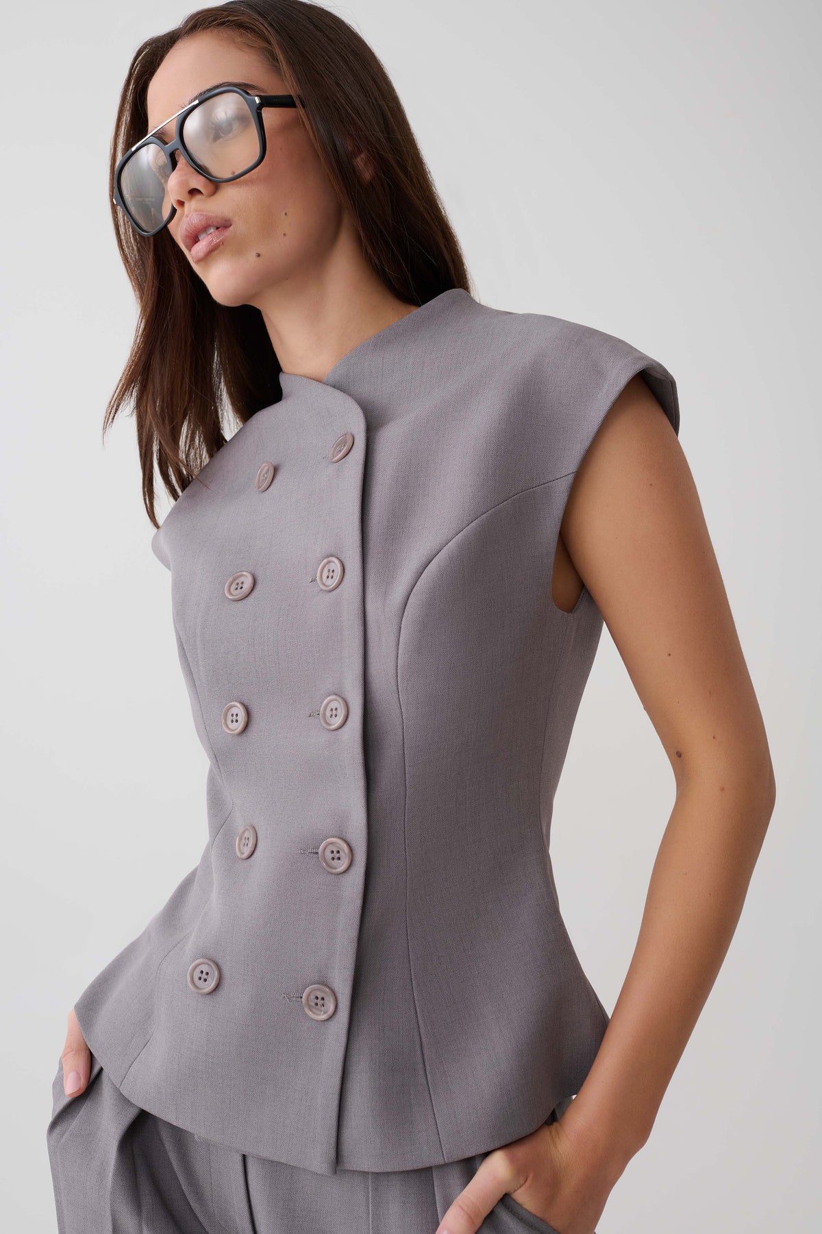 A woman wearing a structured sleeveless peplum-style top in a muted grayish lavender tone, paired with matching trousers and oversized dark sunglasses, against a plain light gray studio background.