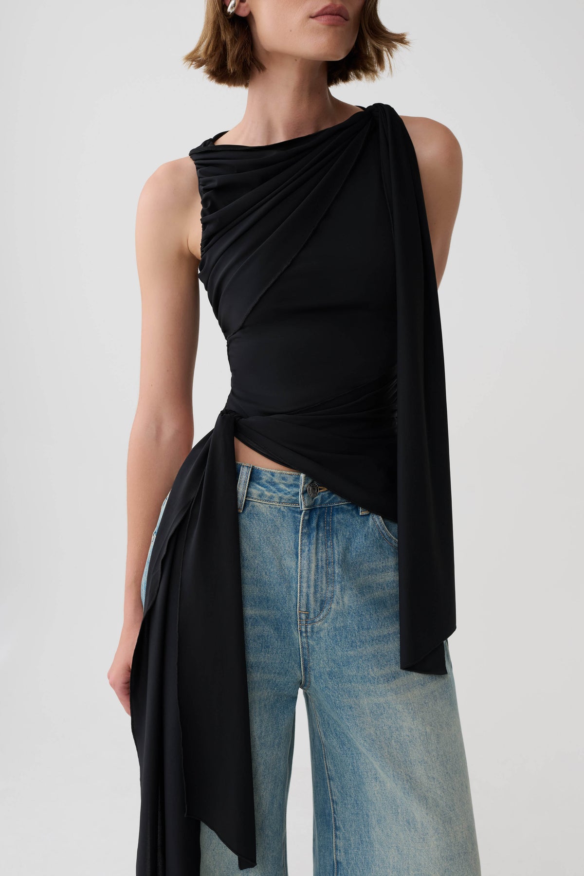 A model wearing a black draped sleeveless top with a side tie detail and light-wash blue jeans, photographed against a light gray studio background.