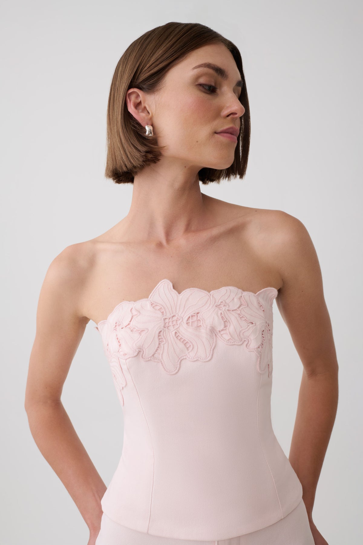 A woman wearing a pale pink strapless corset-style top with floral applique across the bust, photographed against a light gray studio background.