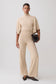 A model stands against a light gray studio background wearing a matching beige knit two-piece set consisting of a short-sleeve mock-neck top and wide-leg trousers, paired with dark brown pointed flats and a small black textured clutch.