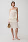 A woman models a cream-colored tiered top with thin spaghetti straps and subtle vertical stripes and matching midi skirt, styled with a small woven straw bucket bag and white slide sandals against a light gray studio background.
