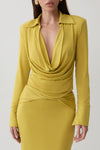 A woman wearing a fitted long-sleeve dress in a rich mustard yellow. The dress features a draped cowl neckline, a cinched waist, and a deep V-neckline.