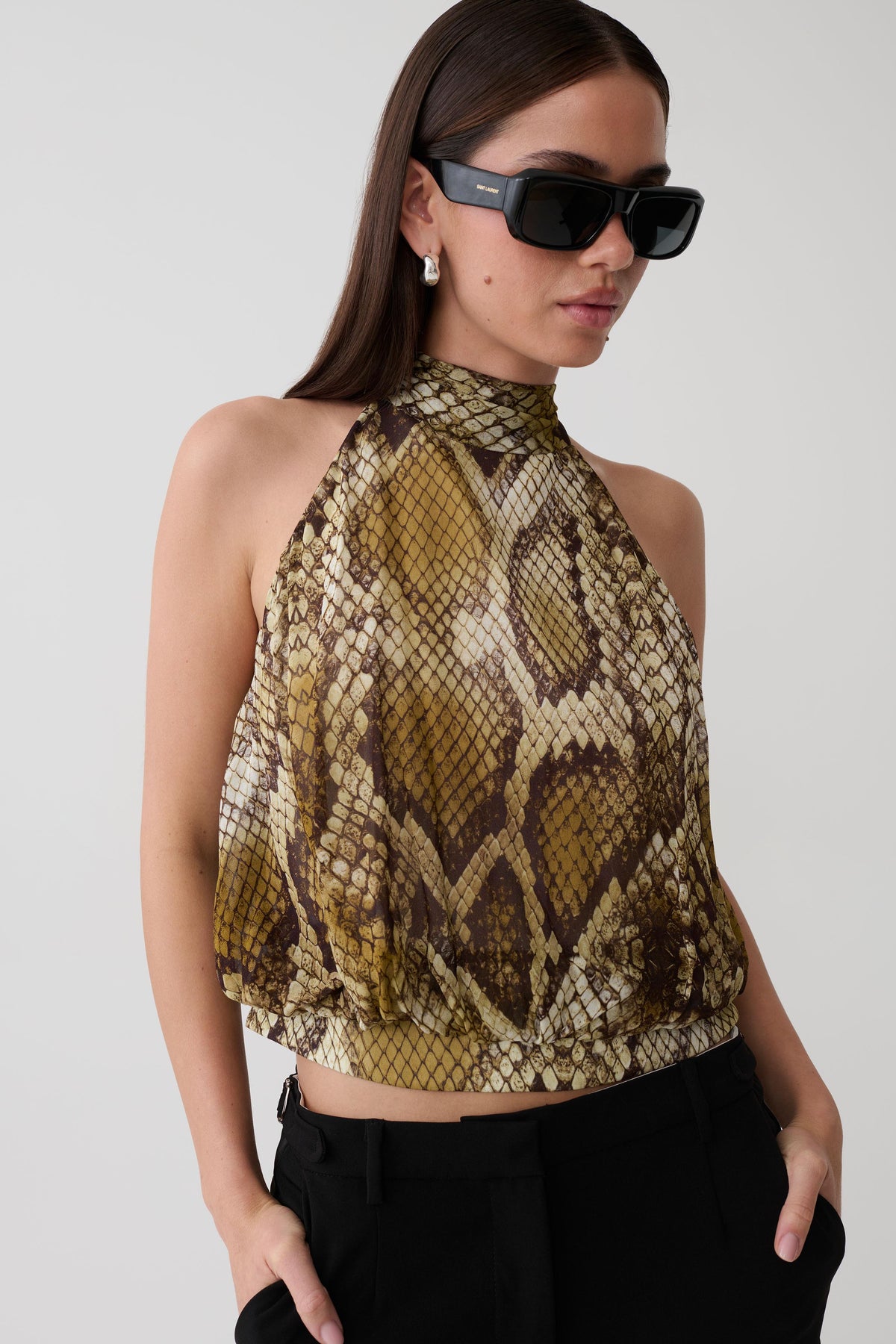 A woman wearing a snakeskin print halter crop top in brown, beige, and black, paired with black pants and black sunglasses, against a light gray studio background.