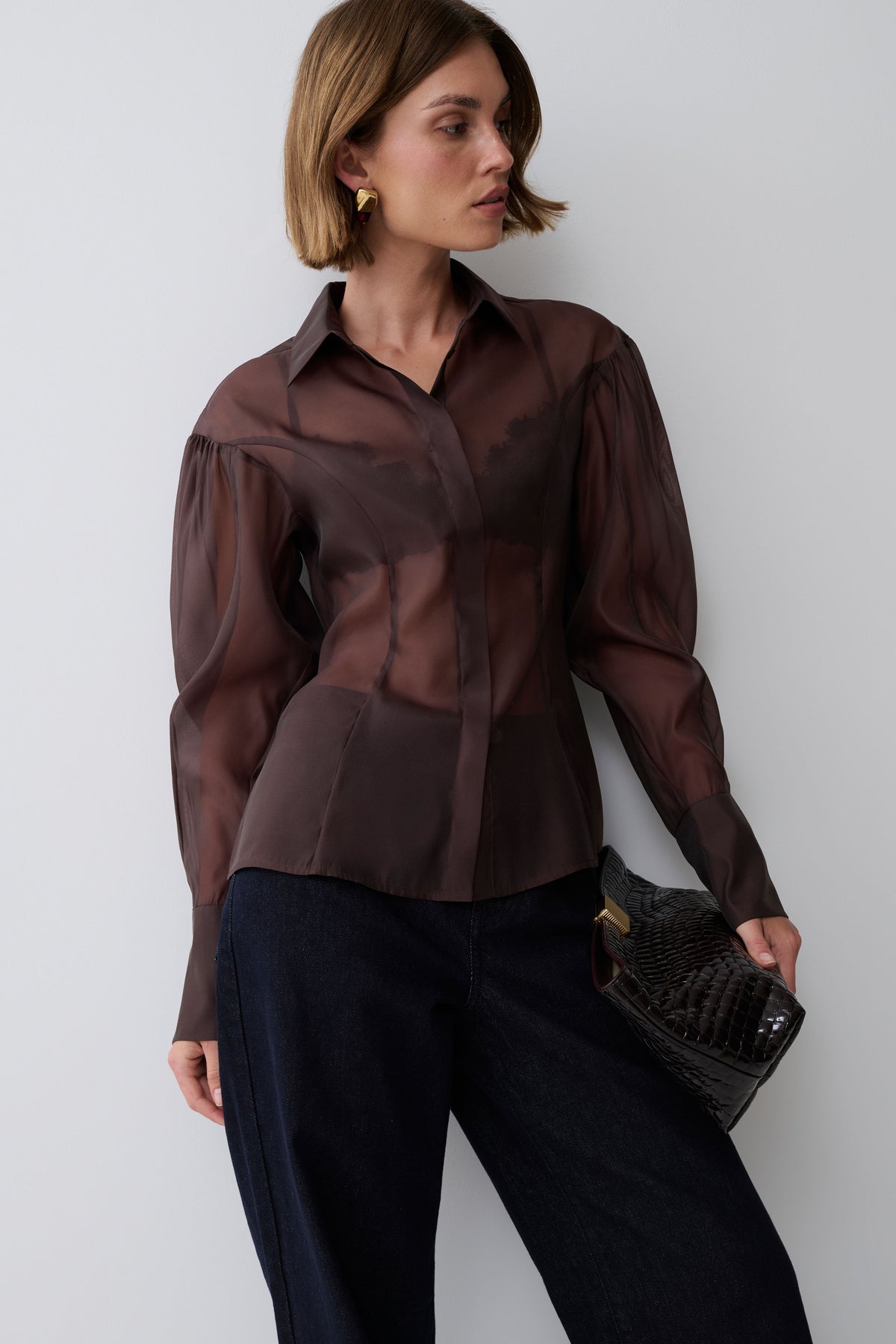 A woman models a sheer, long-sleeved button-up blouse in a dark brown color, paired with dark wide-leg trousers and holding a black textured clutch bag against a plain light gray background.