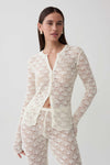 A woman modeling a matching cream-colored crochet knit cardigan and pants set against a plain light gray studio background.