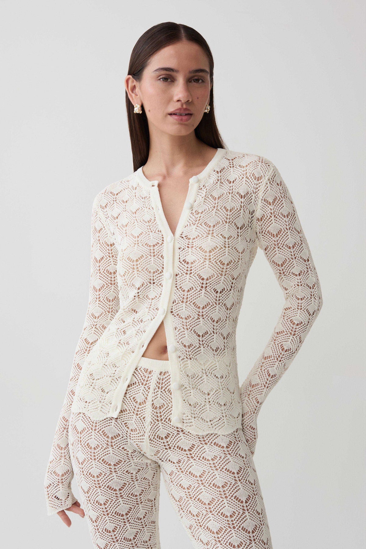 A woman modeling a matching cream-colored crochet knit cardigan and pants set against a plain light gray studio background.