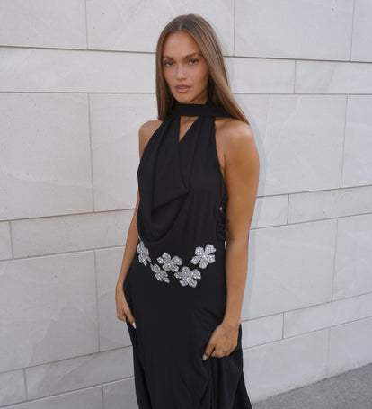 Woman wearing a black halter neck dress with floral embellishments against a light stone wall.