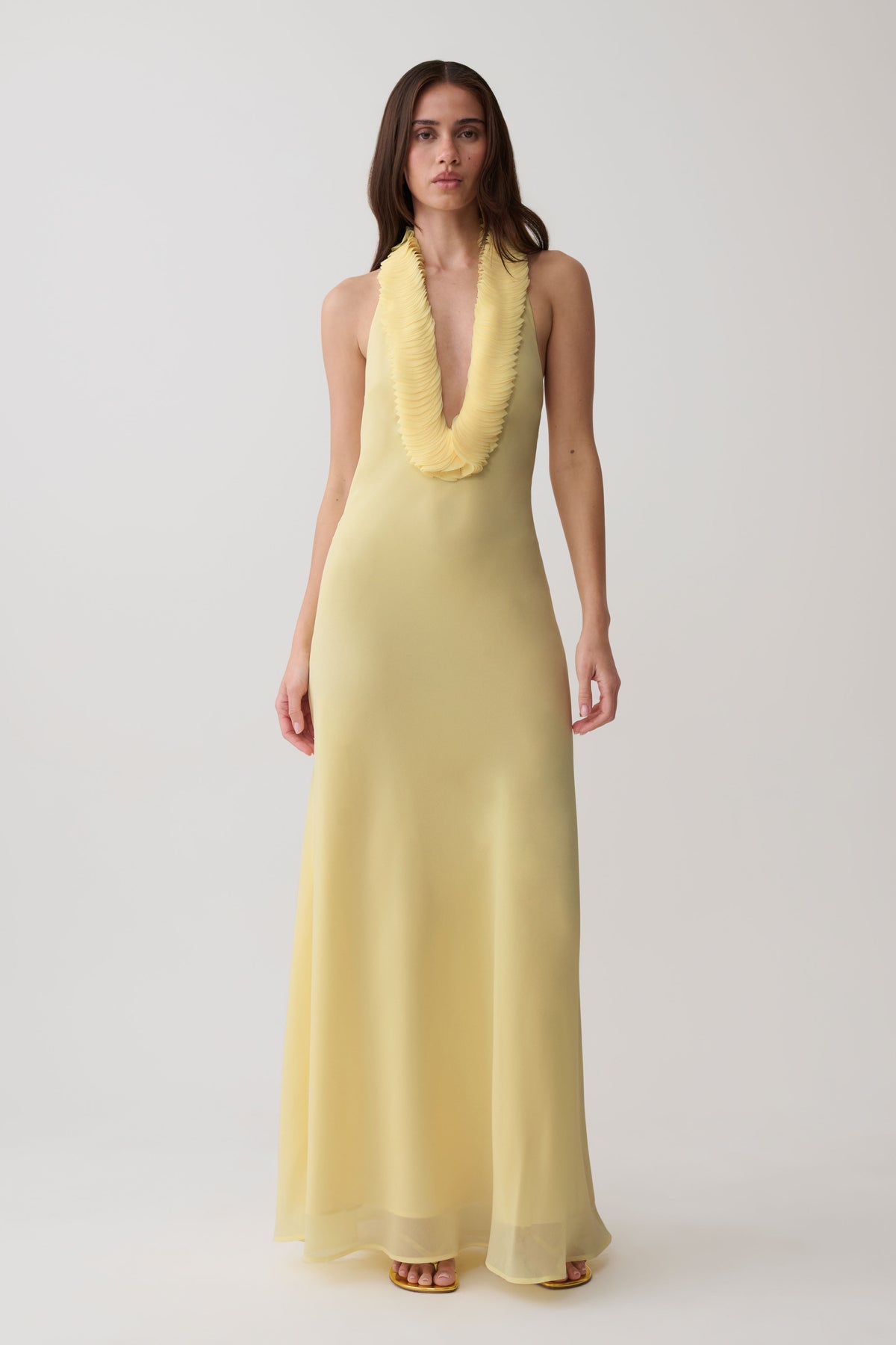 A woman modeling a sleeveless pale yellow maxi dress with a deep V-neckline and a textured, ruffled collar, set against a plain light gray studio background.