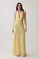 A woman modeling a sleeveless pale yellow maxi dress with a deep V-neckline and a textured, ruffled collar, set against a plain light gray studio background.
