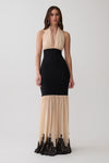 A woman modeling a sleeveless halter-neck maxi dress with a black fitted waist and skirt, and a sheer beige lower skirt with black lace trim at the hem, against a light gray studio background.