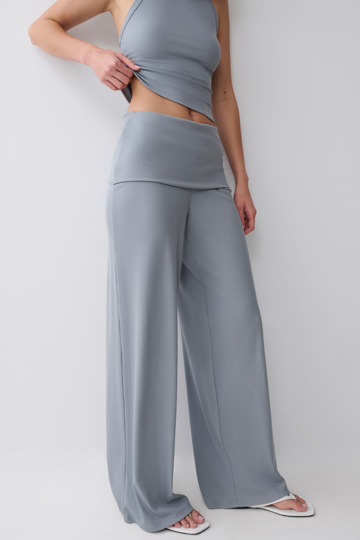 A model wearing a matching two-piece loungewear set consisting of a halter crop top and high-waisted, wide-leg pants in a solid light gray-blue color, styled with white slide sandals against a plain light gray studio background.