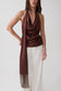 A woman modeling a dark brown draped halter top with a matching long scarf draped over her shoulder, paired with white wide-leg pants, against a plain light gray studio background.