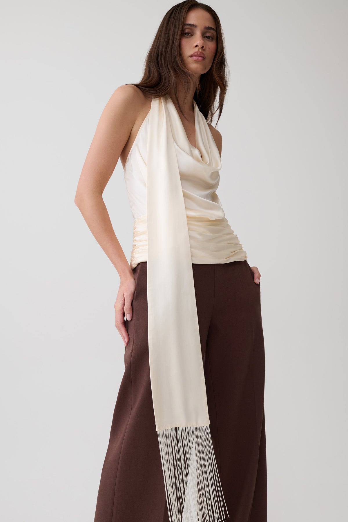 A woman modeling a cream draped halter top with a long cream scarf featuring fringe, paired with dark brown wide-leg pants, against a light gray studio background.