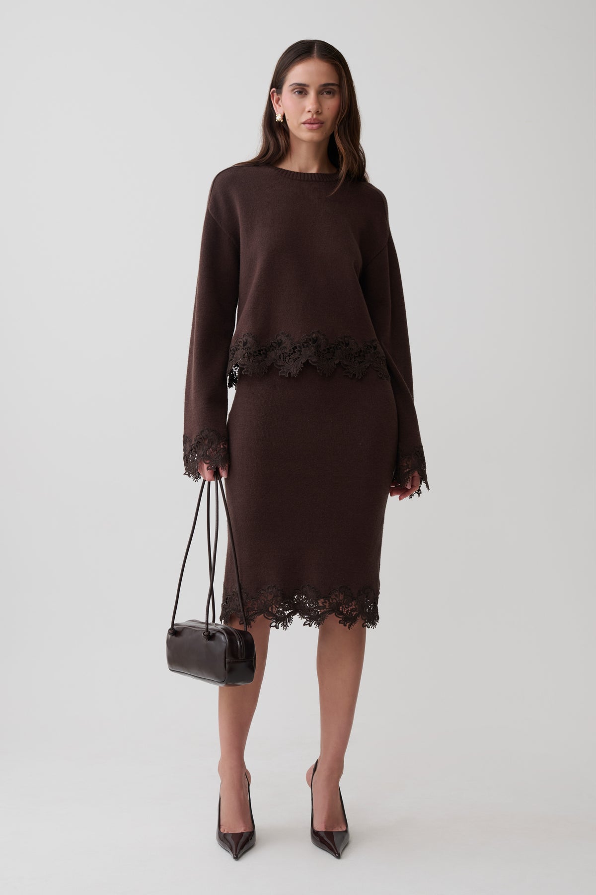 A woman models a dark brown long-sleeve knit jumper and midi skirt with dark brown lace trim, holding a small black handbag against a light gray studio background.
