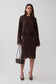 A woman models a dark brown long-sleeve knit jumper and midi skirt with dark brown lace trim, holding a small black handbag against a light gray studio background.